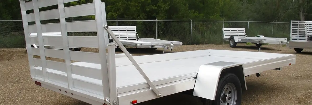White aluminum utility trailer with side ramps and flat deck displayed on gravel lot with additional trailers in the background.